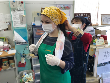 Mary, wearing a face mask and green apron has a yellow headscarf tied for her by a Takahashi Tea colleague. They are stood in the Takahashi Tea office
